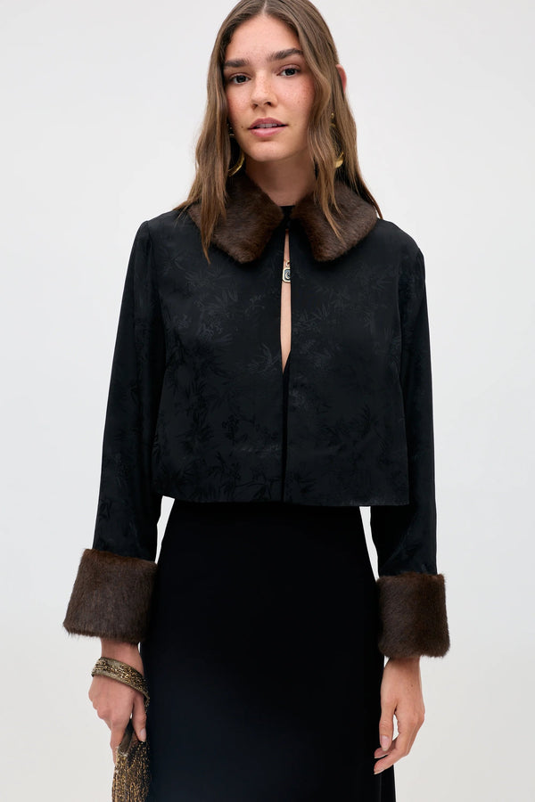 A woman wearing the Luanne - Black jacket, featuring a subtle floral pattern, brown faux fur collar and cuffs, paired with a black skirt, stands against a light background.