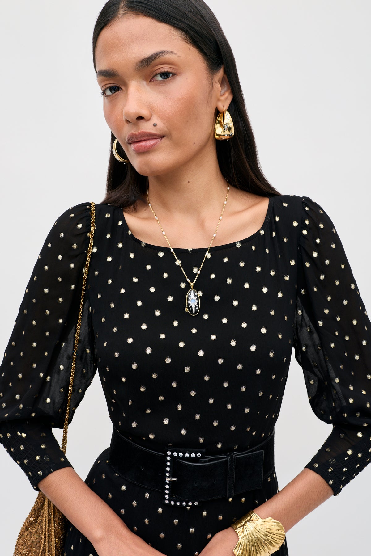 A woman with long dark hair wears the “Eileen - Black” dress, featuring gold polka dots, paired with gold earrings, a cameo necklace, a black belt with a gold buckle, a gold leaf-shaped bracelet, and carries a matching gold purse.