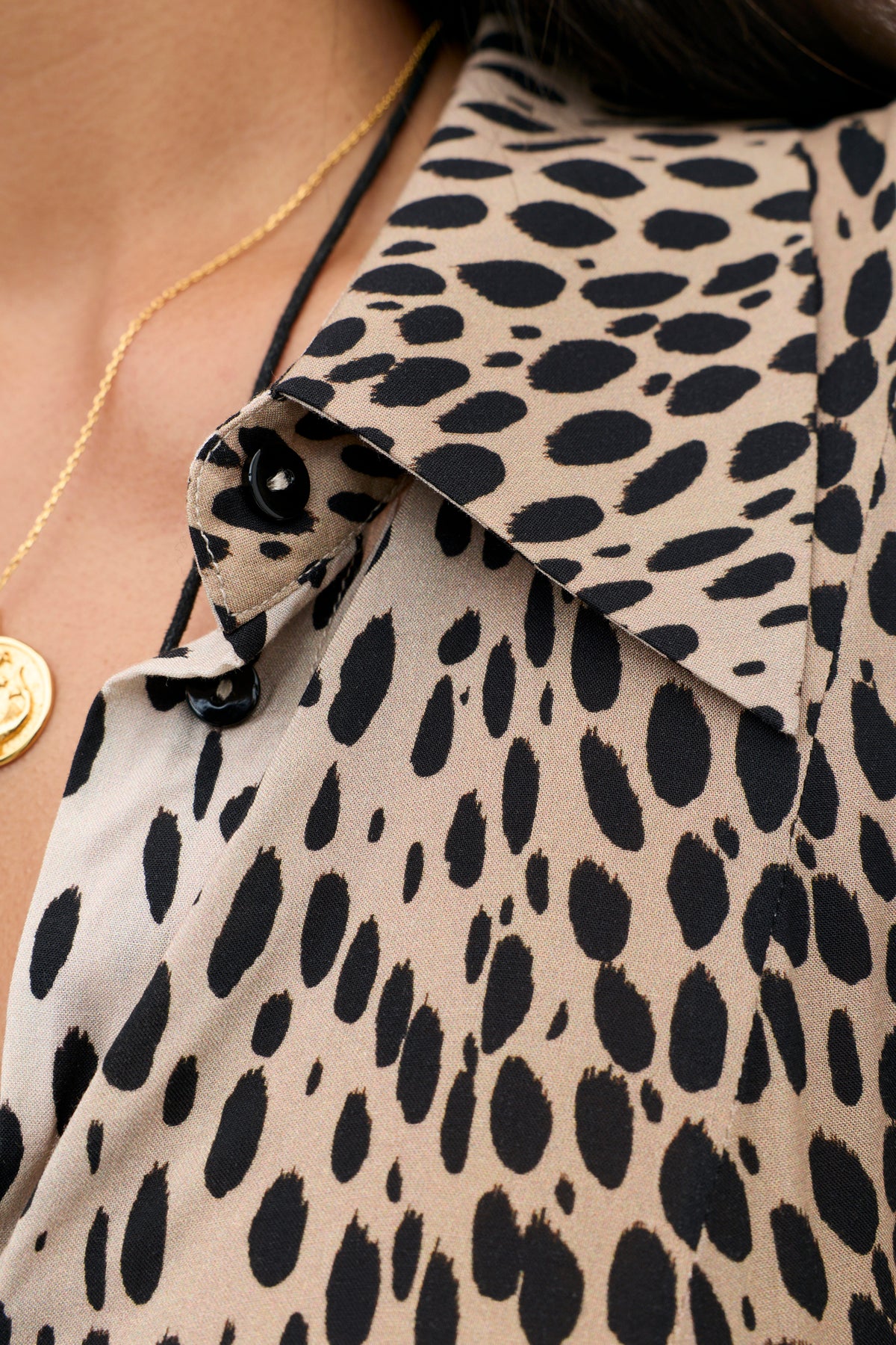 Close-up of the Maddison - Antelope Spot Black: a beige shirt dress with black leopard spots, black buttons, and a gold pendant necklace on a petite person—perfect for elevating your everyday styles.