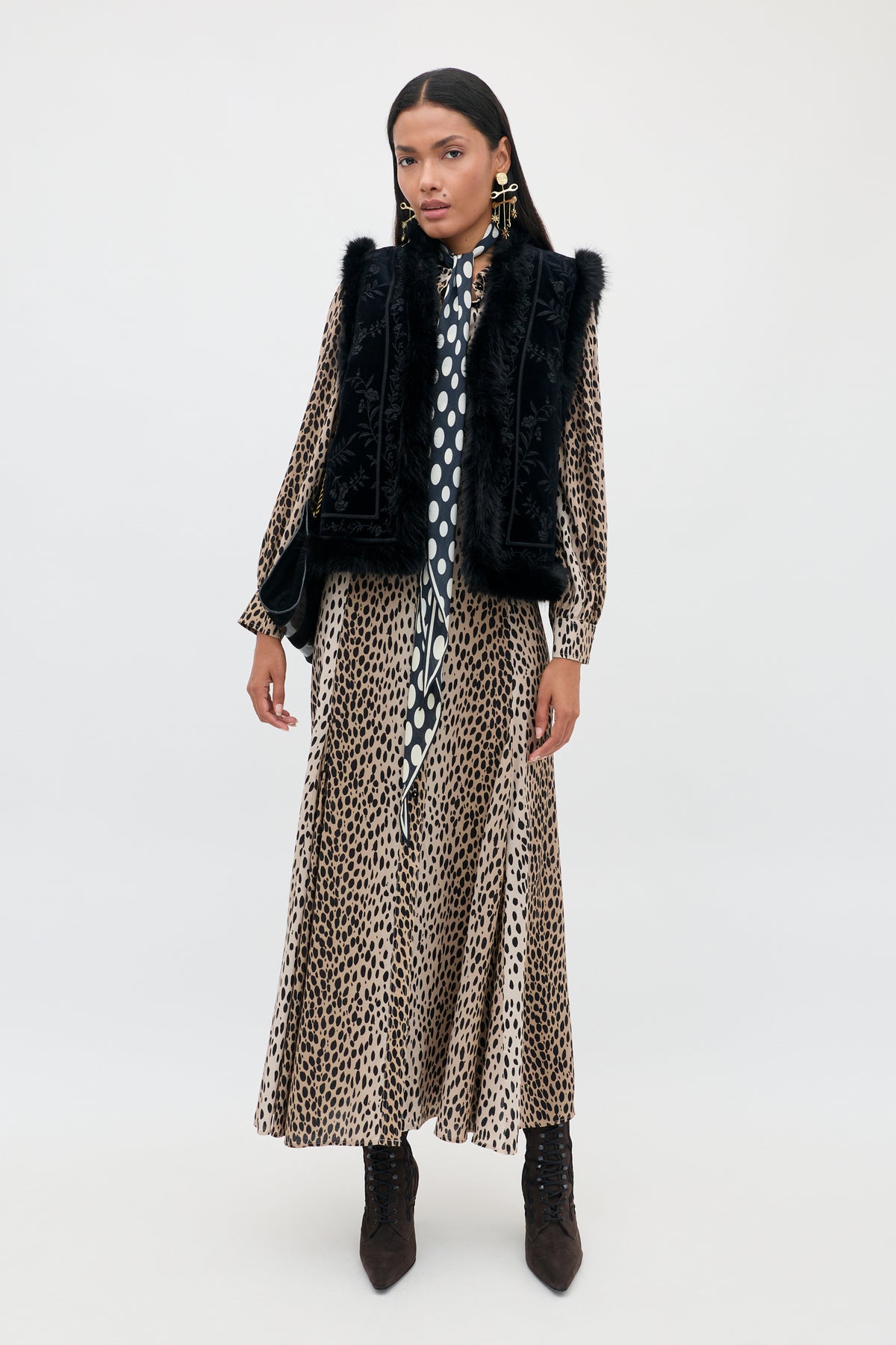 A model wears a leopard print midi dress with a polka dot necktie, a Taletha - Botanical Vine Embroidery Black vest with faux fur trim, and dark brown ankle boots.