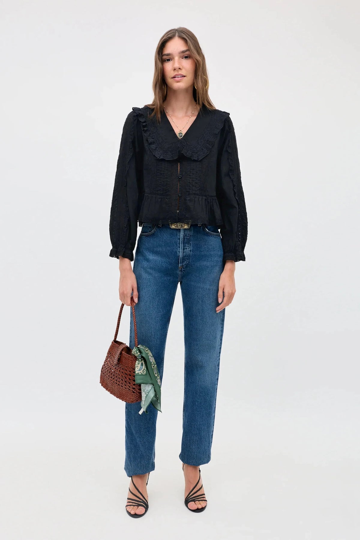 A woman wears a relaxed fit, long-sleeved Maribelle - Black blouse with a wide collar, blue jeans, and strappy black heels, holding a small brown woven handbag with a green patterned scarf attached.