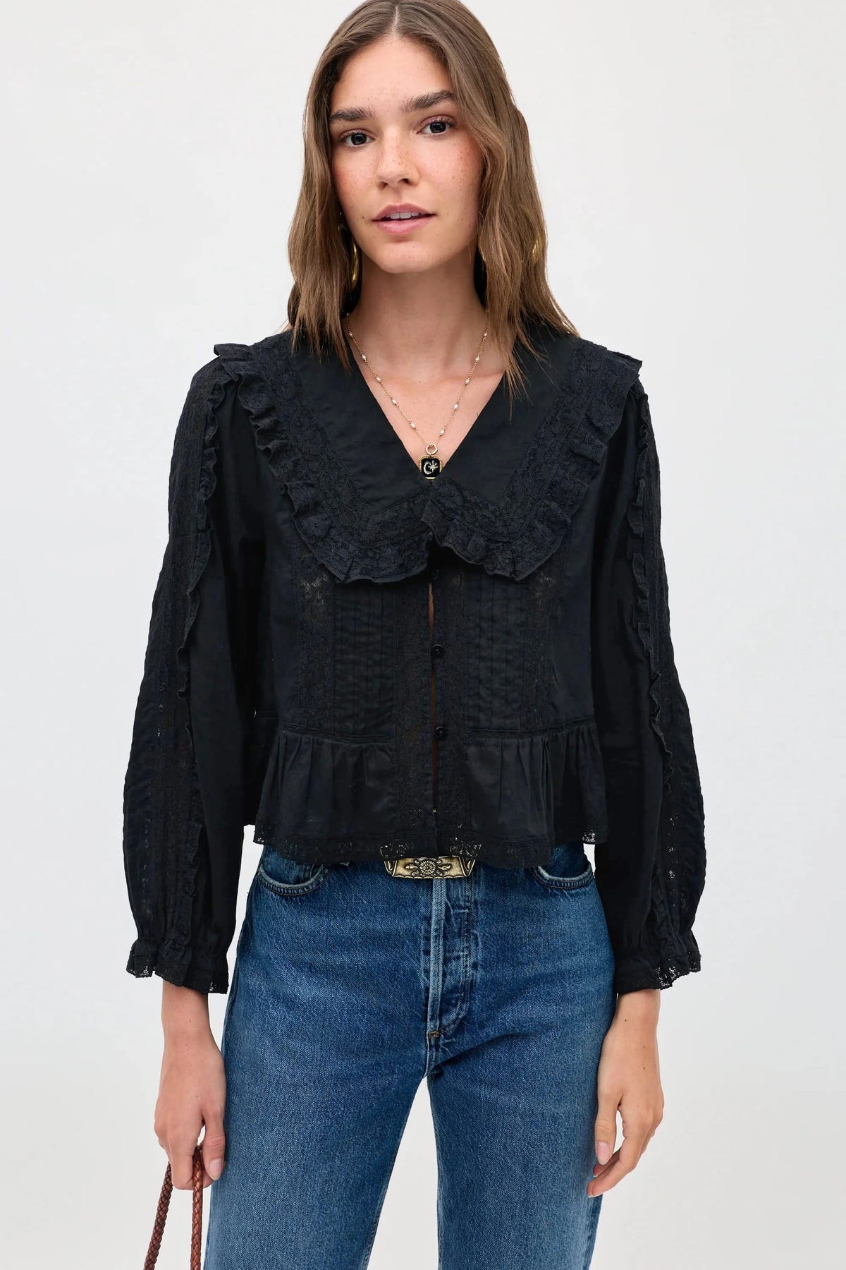 A woman wearing the Maribelle - Black, a vintage-inspired black blouse with a large lace-trimmed collar, button front, and a ruffled hem, styled in a relaxed fit with blue jeans.
