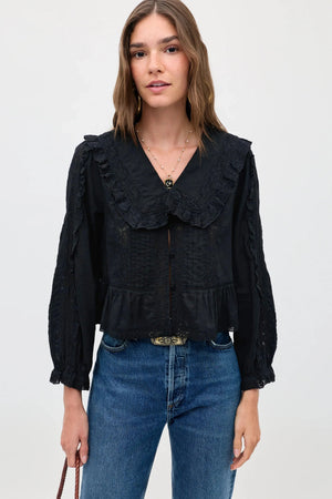 A woman wearing the Maribelle - Black, a vintage-inspired black blouse with a large lace-trimmed collar, button front, and a ruffled hem, styled in a relaxed fit with blue jeans.