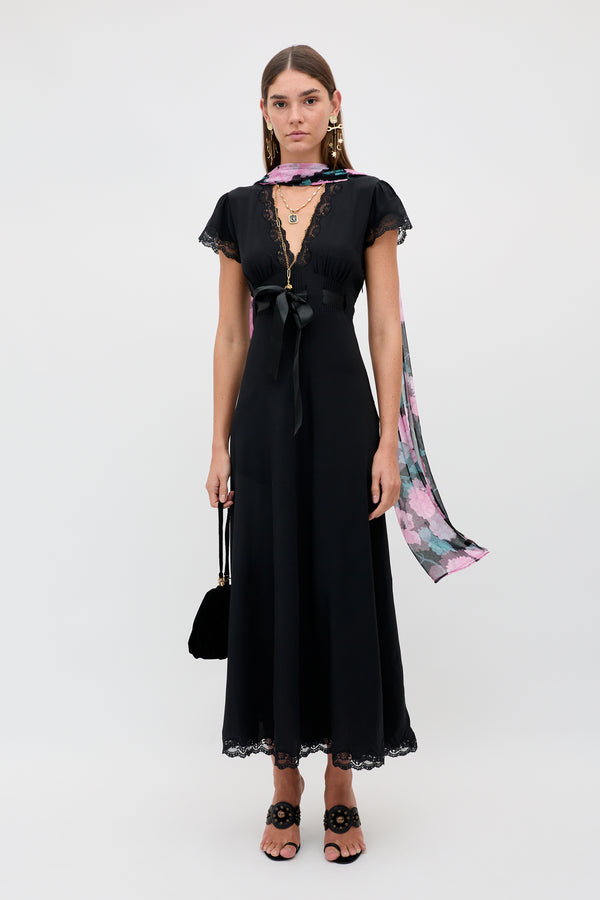 A woman stands against a plain white background wearing the Marlice - Black: a black, short-sleeved, ankle-length dress with lace trim. She accessorizes her look with statement earrings, a pink floral scarf tied in a delicate satin bow, black heeled sandals, and carries a coordinating black handbag.