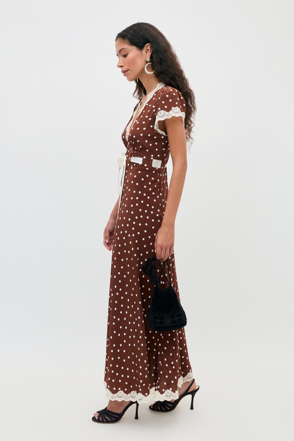A woman wearing the Marlice - Layla Dot Brown maxi dress, featuring short sleeves, polka-dot pattern, and white lace trim. She is accessorized with a small black handbag, black heeled sandals, and large hoop earrings, standing against a plain background.