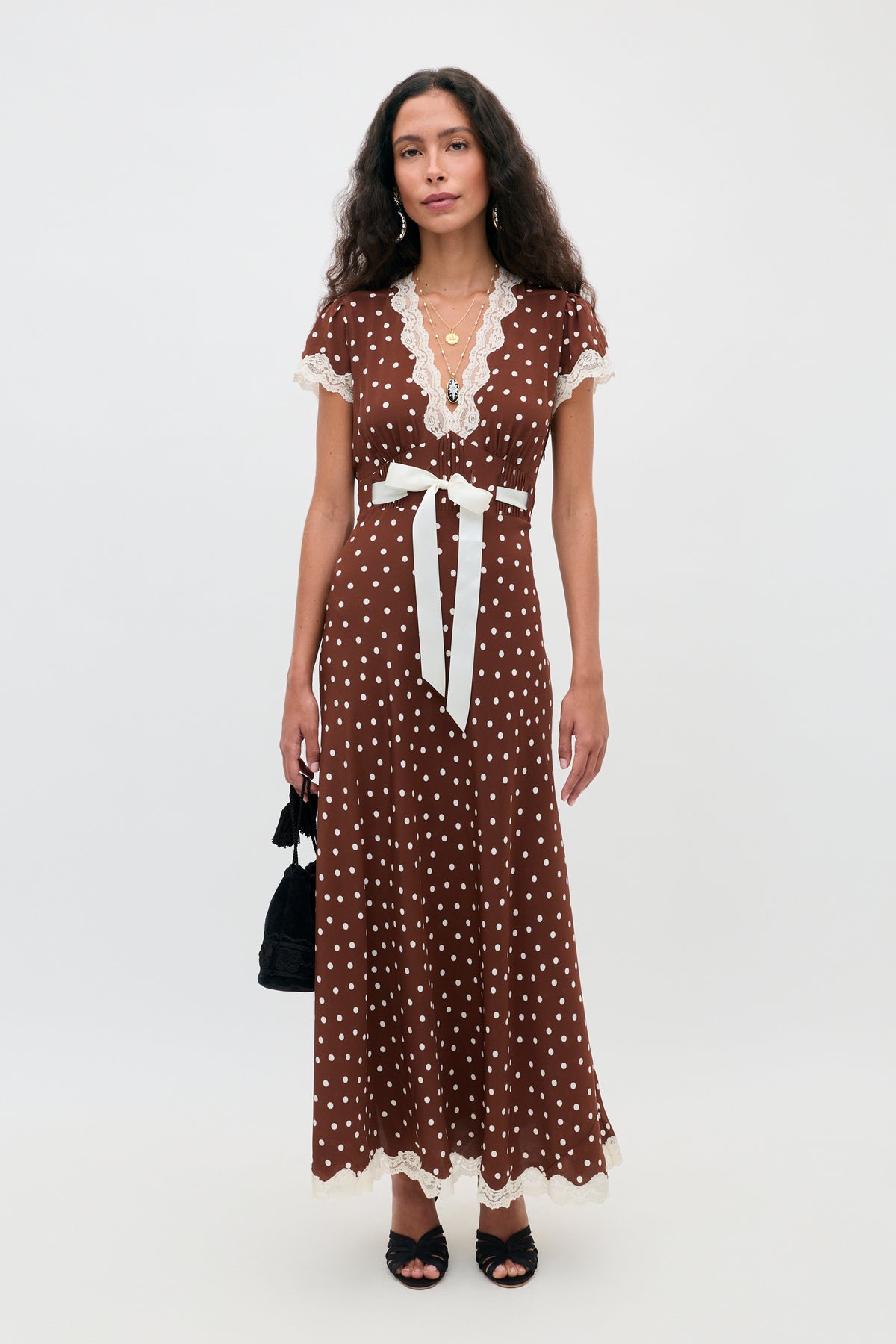 A woman wearing the Marlice - Layla Dot Brown maxi dress, featuring short sleeves, polka dots, lace trim, and a white waist tie, stands against a plain background while holding a small black handbag and wearing black heels.
