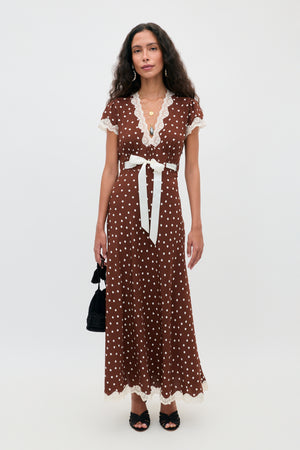 A woman wearing the Marlice - Layla Dot Brown maxi dress, featuring short sleeves, polka dots, lace trim, and a white waist tie, stands against a plain background while holding a small black handbag and wearing black heels.