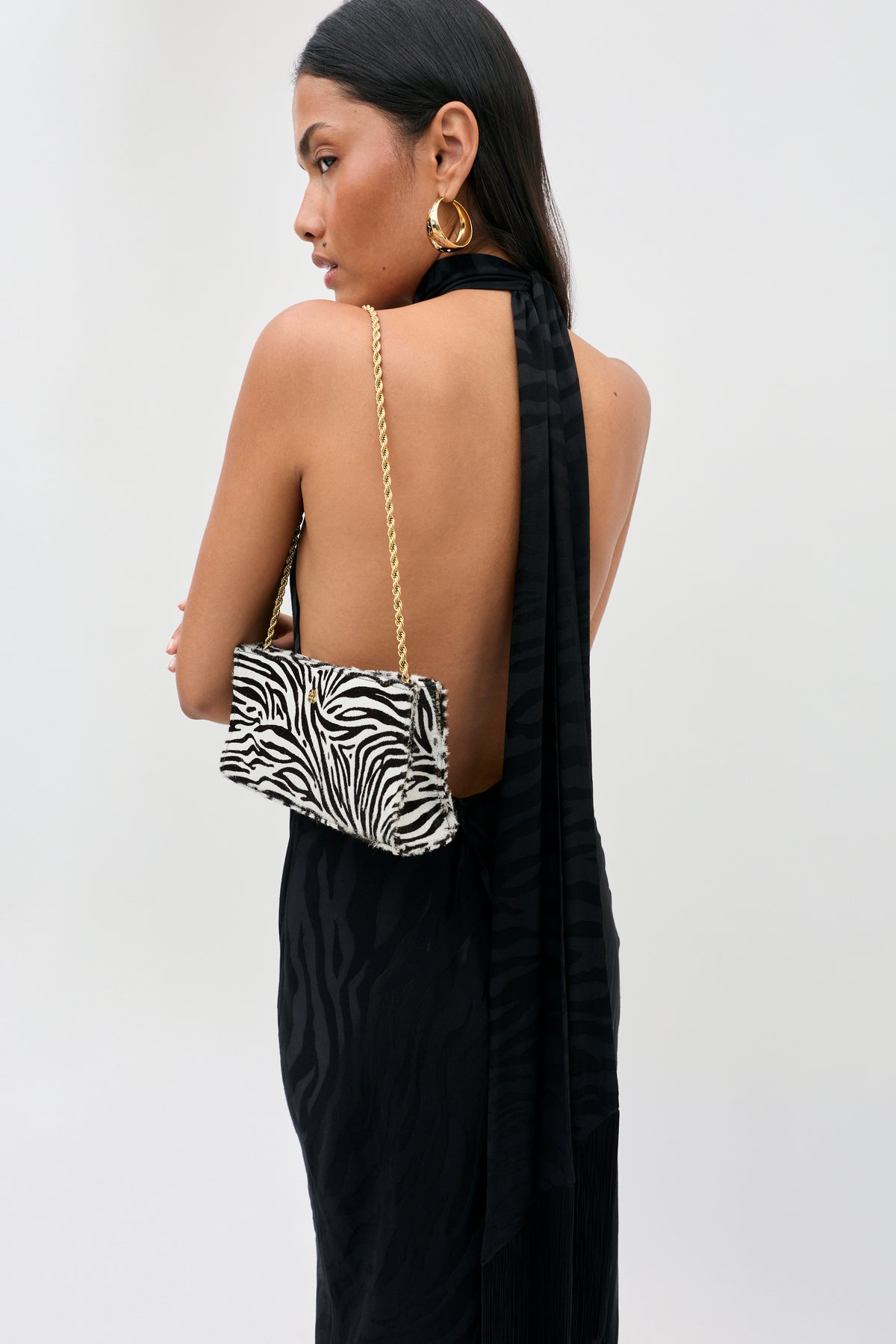 A person wearing a backless black dress carries a Kamila - Zebra Print Brown shoulder bag featuring a brown and white zebra print and a gold chain strap.