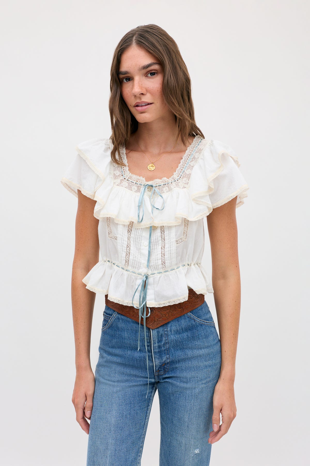 Woman with long brown hair wearing the melisa top ivory—a prairie-inspired white cotton blouse featuring lace ruffles and blue ribbon accents—along with blue jeans and a brown belt, standing against a plain light background.