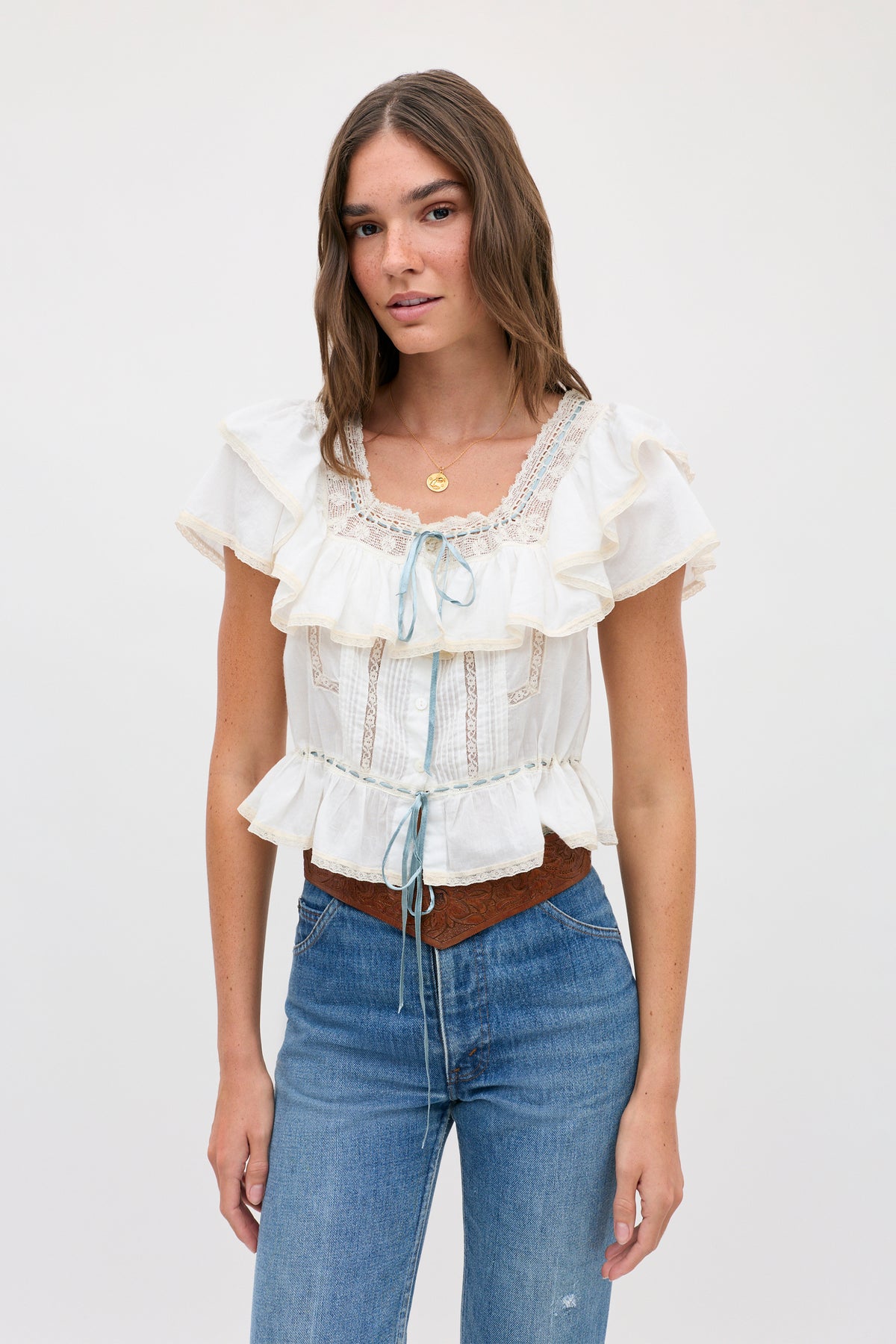 Woman with long brown hair wearing the melisa top ivory—a prairie-inspired white cotton blouse featuring lace ruffles and blue ribbon accents—along with blue jeans and a brown belt, standing against a plain light background.