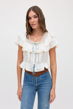 Woman with long brown hair wearing the melisa top ivory—a prairie-inspired white cotton blouse featuring lace ruffles and blue ribbon accents—along with blue jeans and a brown belt, standing against a plain light background.