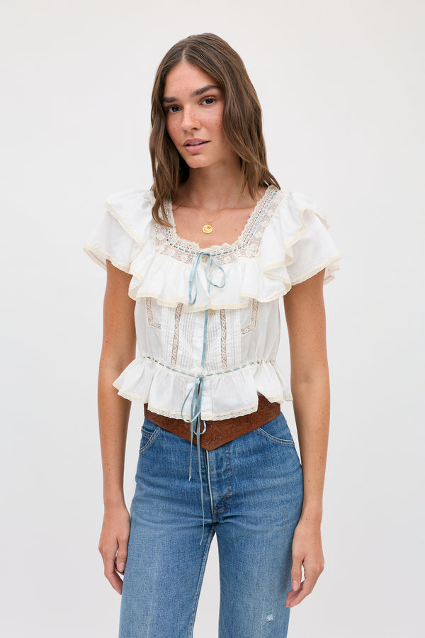 Woman with long brown hair wearing the melisa top ivory—a prairie-inspired white cotton blouse featuring lace ruffles and blue ribbon accents—along with blue jeans and a brown belt, standing against a plain light background.