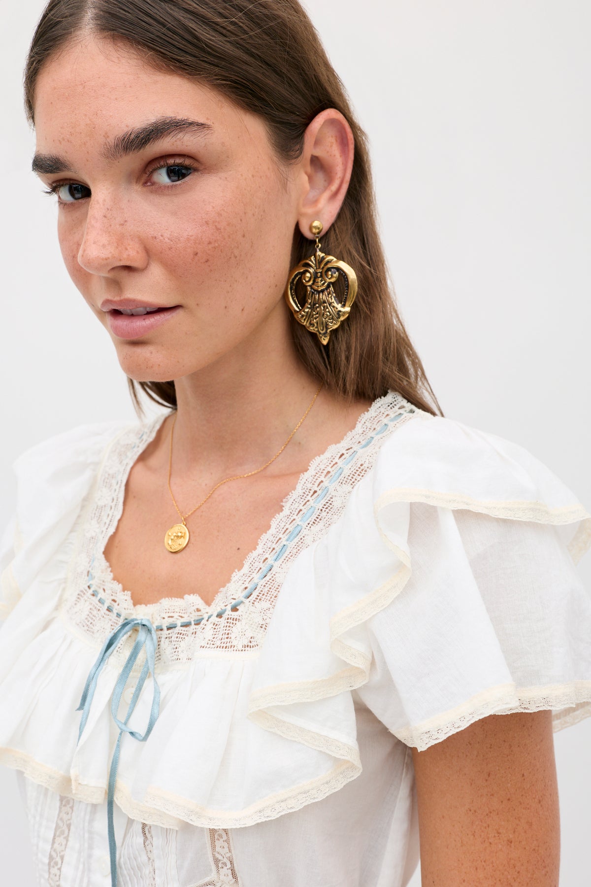 A young woman with long brown hair wears the melisa top ivory, a prairie-inspired white cotton blouse adorned with ruffles, along with gold earrings and a gold necklace. She has light makeup, freckles, and a slight smile. The background is plain and light-colored.