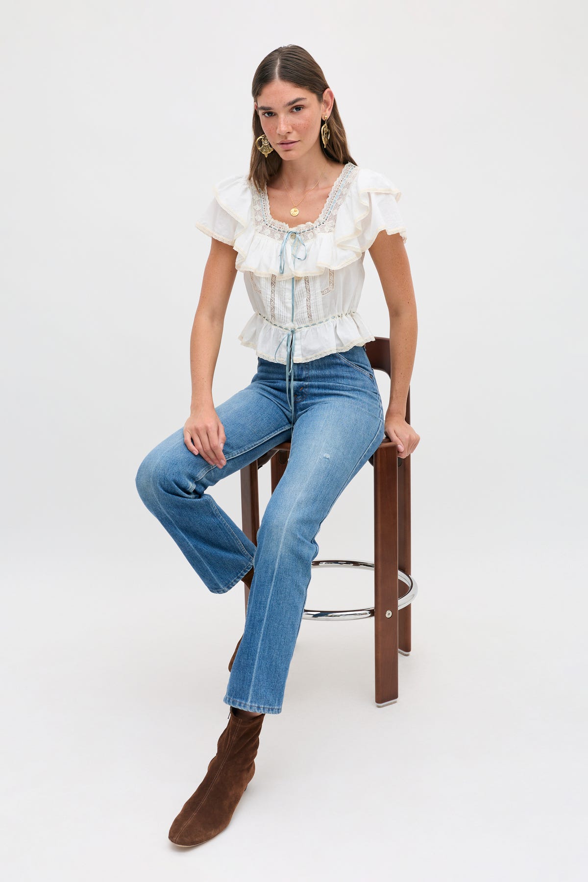 A woman is seated on a wooden stool against a plain white background, wearing the melisa top ivory—a prairie-inspired white cotton blouse adorned with delicate ruffles—paired with blue jeans and brown heeled boots. She has straight brown hair and accessorizes with gold hoop earrings.