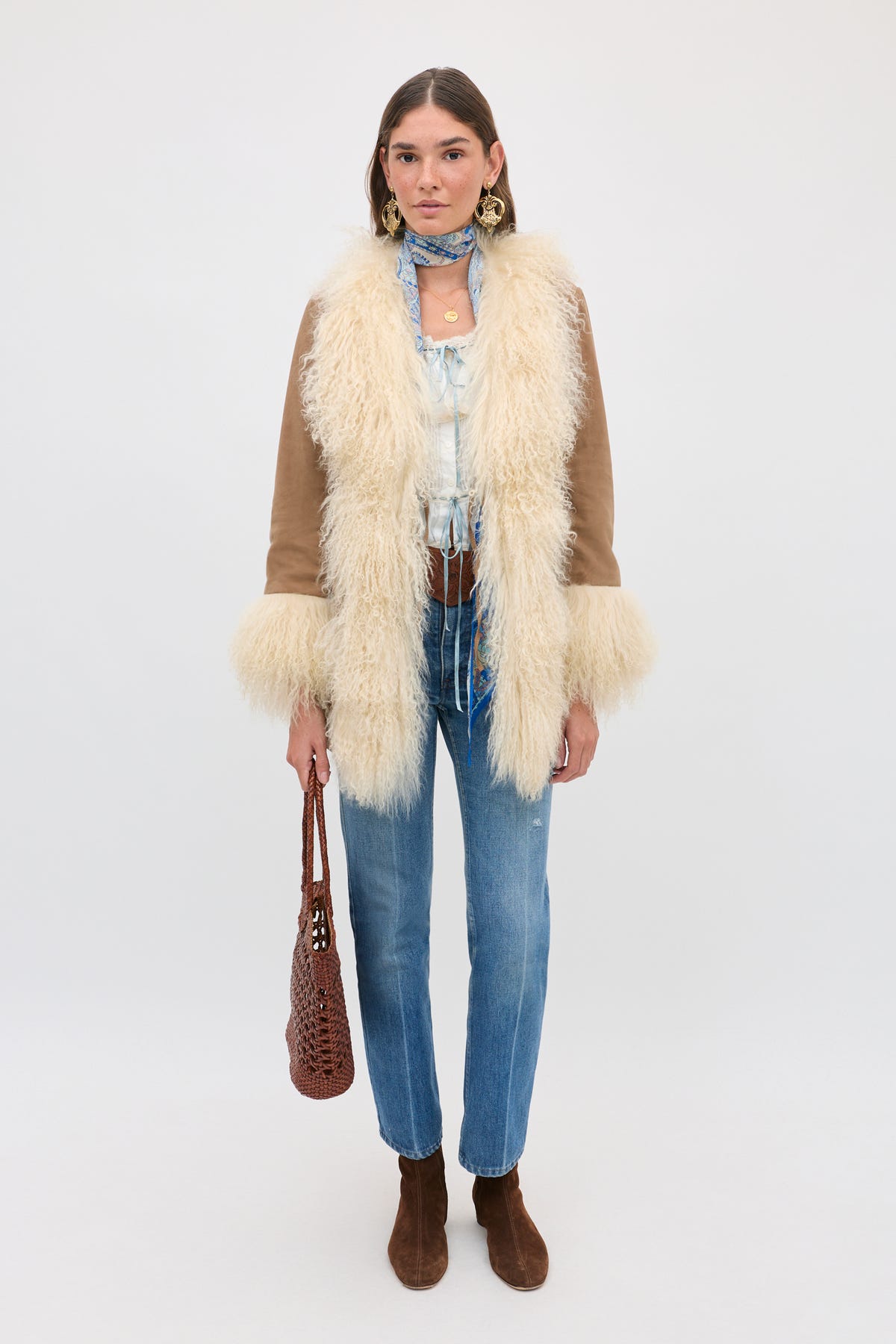 Wearing the melisa top in ivory—a white cotton blouse with delicate ruffles—layered under a tan and cream fur-trimmed coat, this woman pairs her look with jeans, brown boots, a blue scarf, earrings, and carries a brown woven bag, standing against a plain light background for prairie-inspired charm.