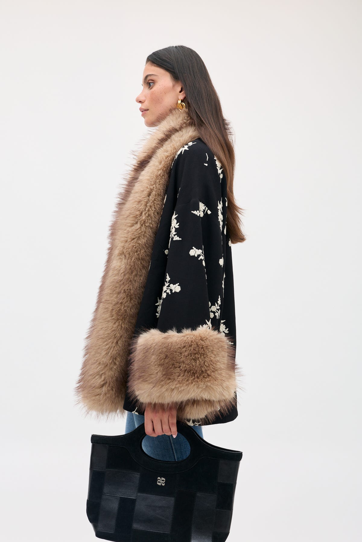 A woman wearing a Micah - Alexa Flower Embroidery Black jacket with cream flower embroidery and brown faux fur trim on the collar and cuffs, holding a black handbag.