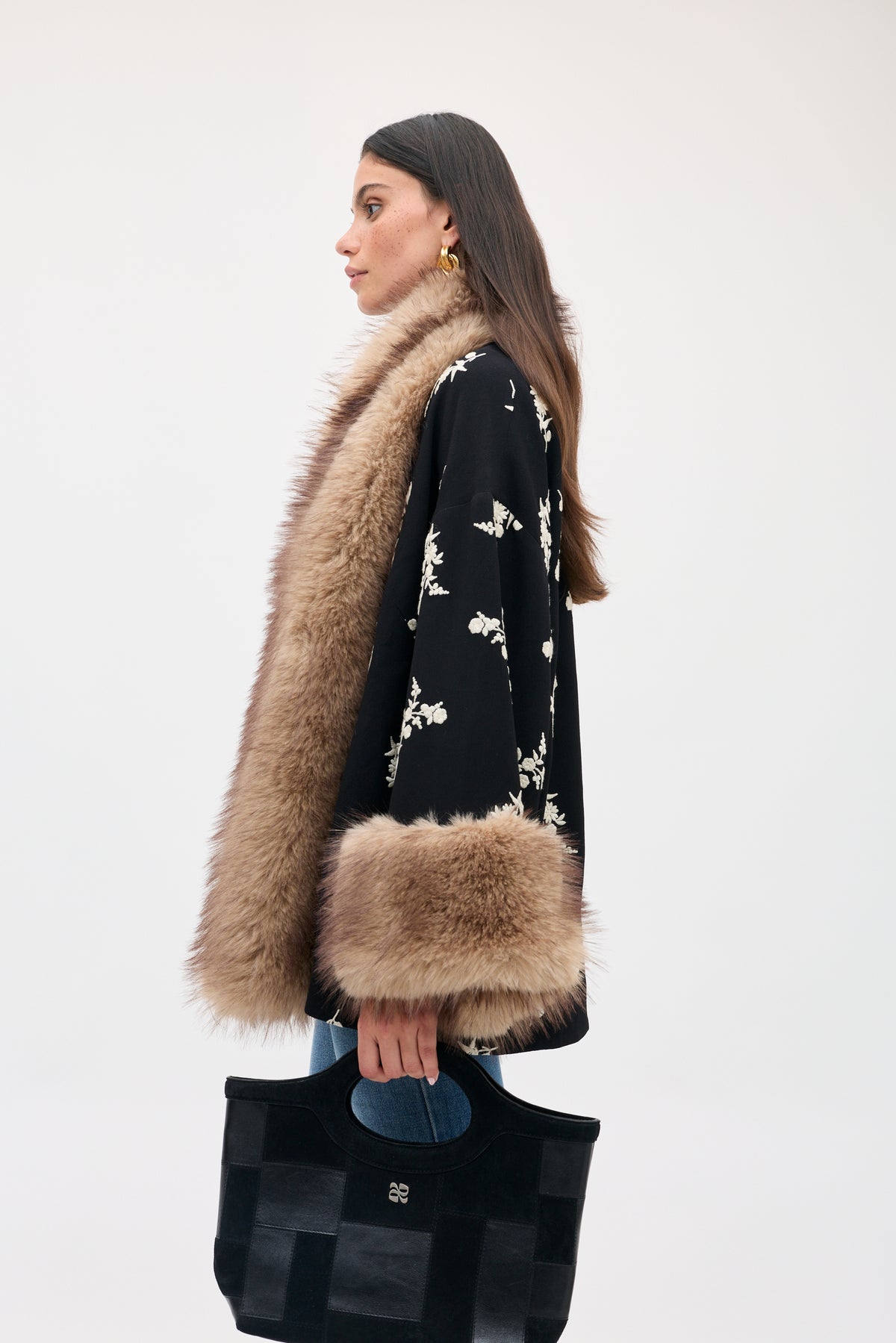 A woman wearing a Micah - Alexa Flower Embroidery Black jacket with cream flower embroidery and brown faux fur trim on the collar and cuffs, holding a black handbag.