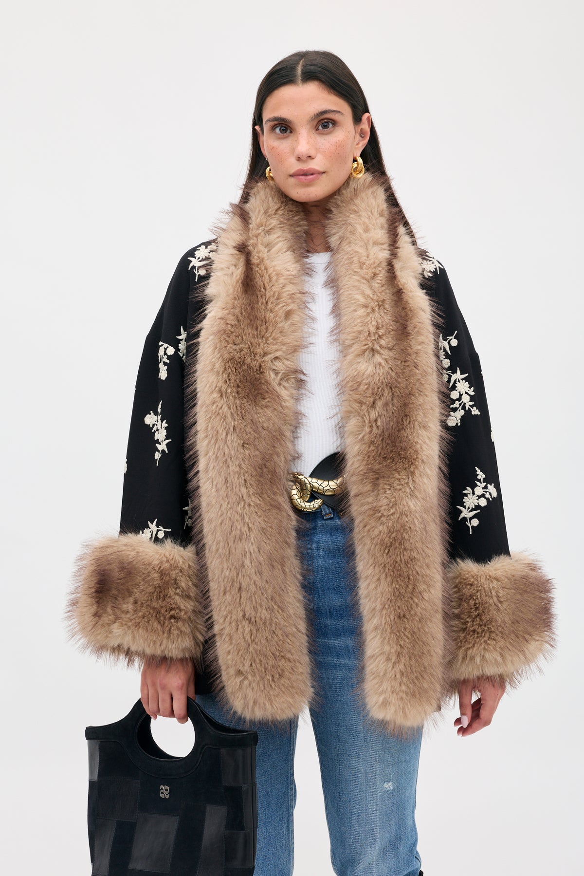 A person wears the Micah - Alexa Flower Embroidery Black coat featuring white floral embroidery and a wide, fluffy faux fur collar and cuffs, paired with blue jeans and a black tote bag.