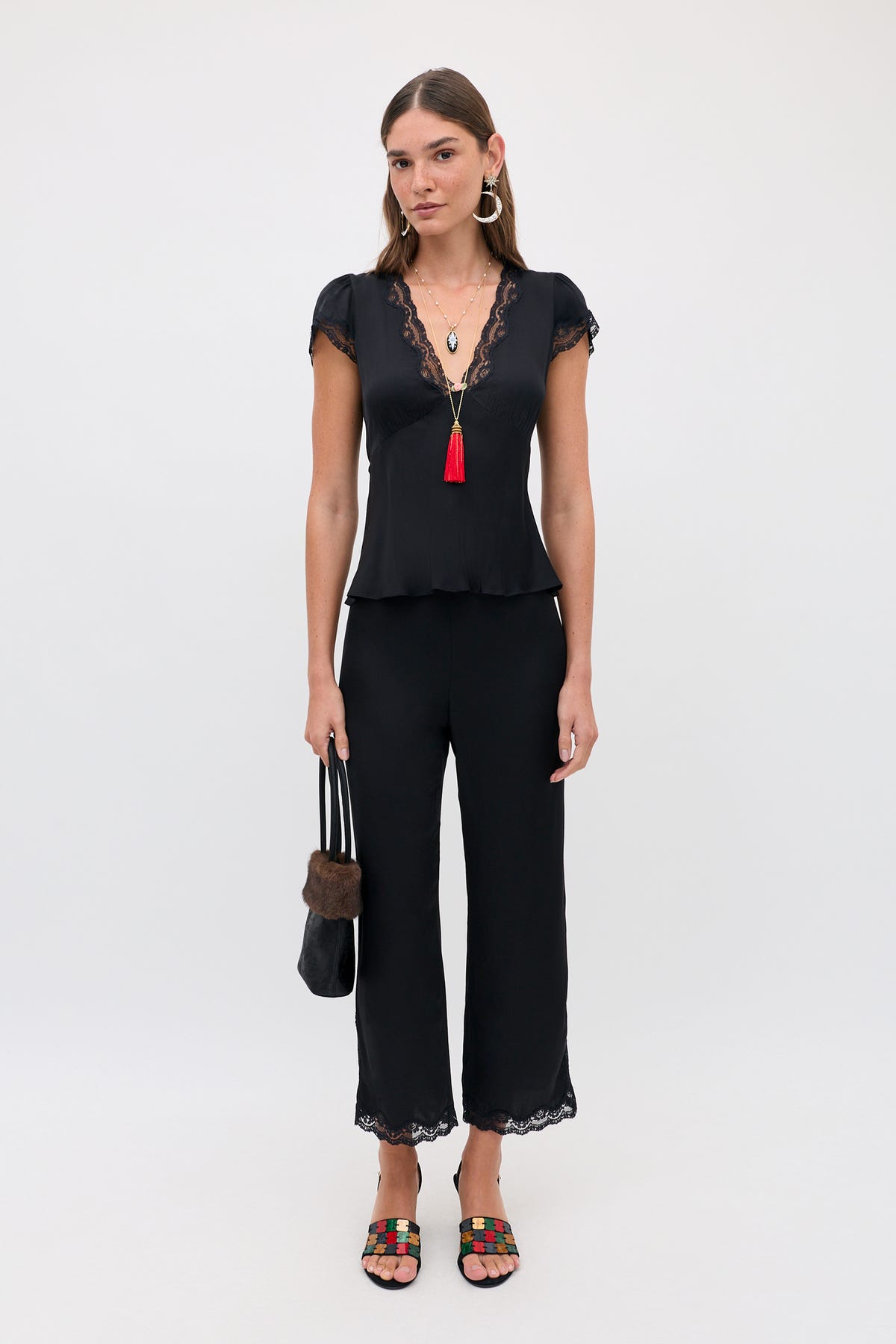 Woman standing against a plain background, wearing the Mireille - Black lace-trimmed top and pants, colorful sandals, hoop earrings, layered necklaces with a red tassel, and holding a black fur-trimmed bag.