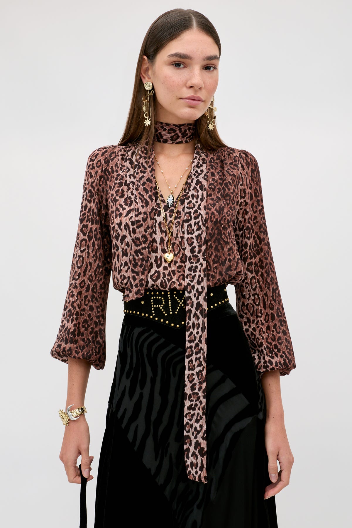 A woman is wearing the Moss - Leopard Brown vintage-inspired silk blouse with a matching scarf, paired with a black textured skirt, gold jewelry, and dangling earrings, standing against a plain light background.