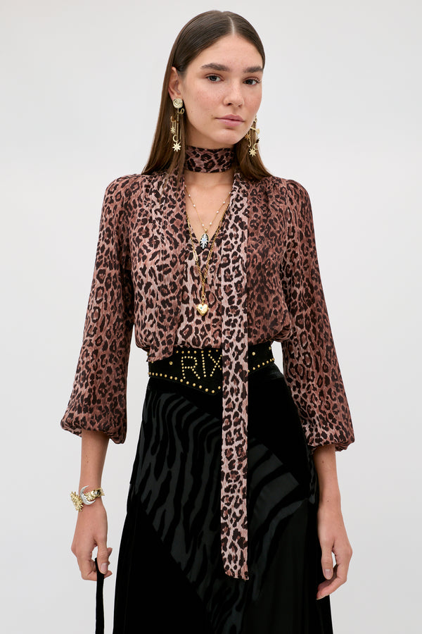 A woman is wearing the Moss - Leopard Brown vintage-inspired silk blouse with a matching scarf, paired with a black textured skirt, gold jewelry, and dangling earrings, standing against a plain light background.