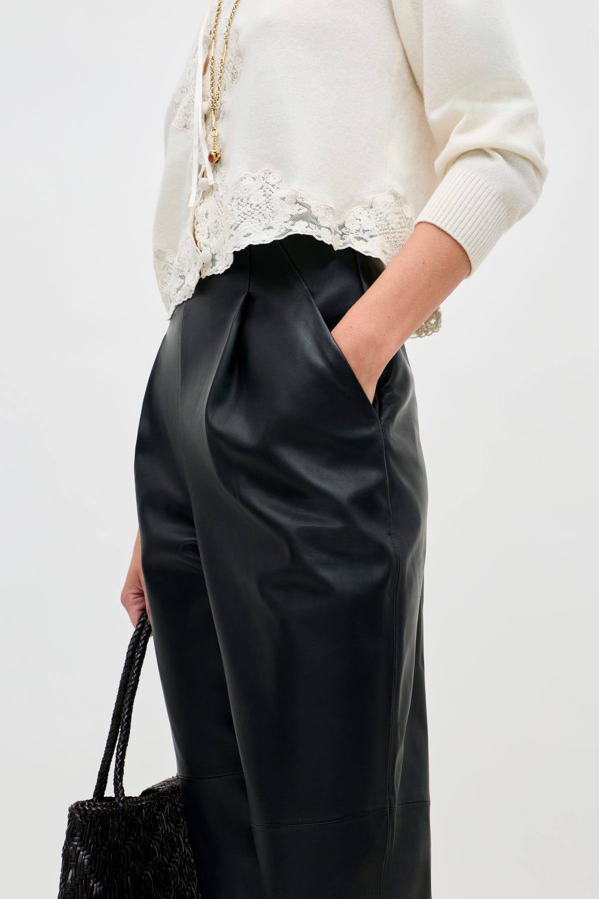 A person wearing the Nori - Charcoal sweater with white lace trim and high-waisted black leather pants, hands in pockets and holding a black woven handbag. The photo is cropped at the shoulders and knees.