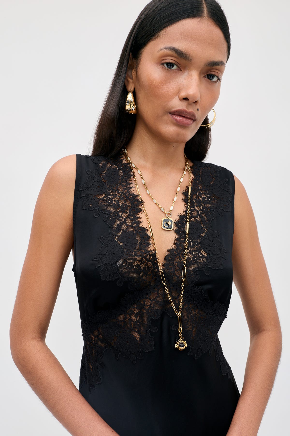 A woman with long dark hair wears the “Rhegan - Black” sleeveless lace dress, layered gold necklaces, and gold earrings. She stands against a plain light background with a neutral expression, looking slightly past the camera.