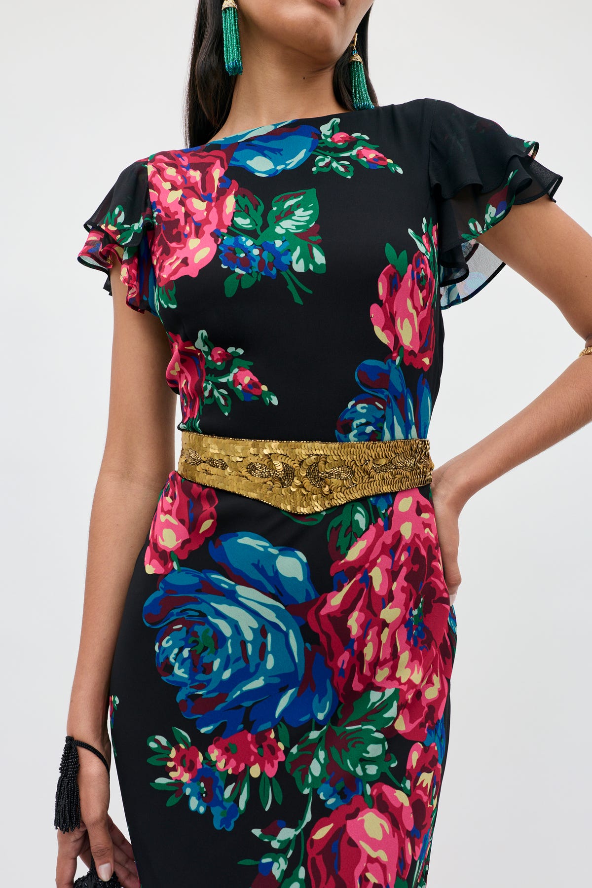 A woman poses with one hand on her hip against a plain background, wearing the Rorie - Vintage Flora Embellishment Antique Gold dress featuring bold floral prints and a gold belt, styled with green earrings and a black bracelet.