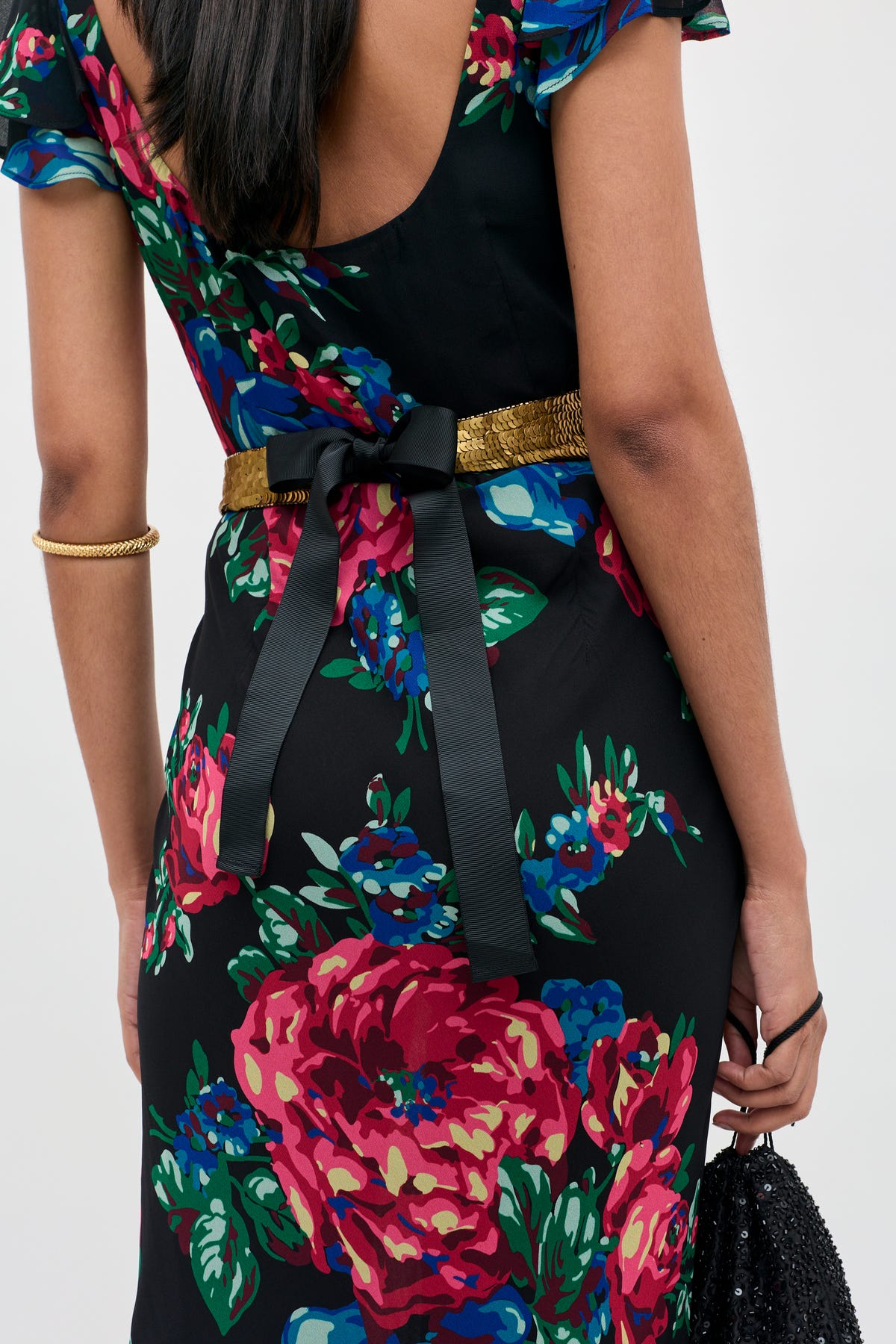 Wearing the Rorie - Vintage Flora Embellishment Antique Gold dress, she accessorizes with a gold bracelet, a gold belt featuring a black bow, and carries a black beaded clutch. The elegant dress showcases a black floral design and a low-back silhouette.