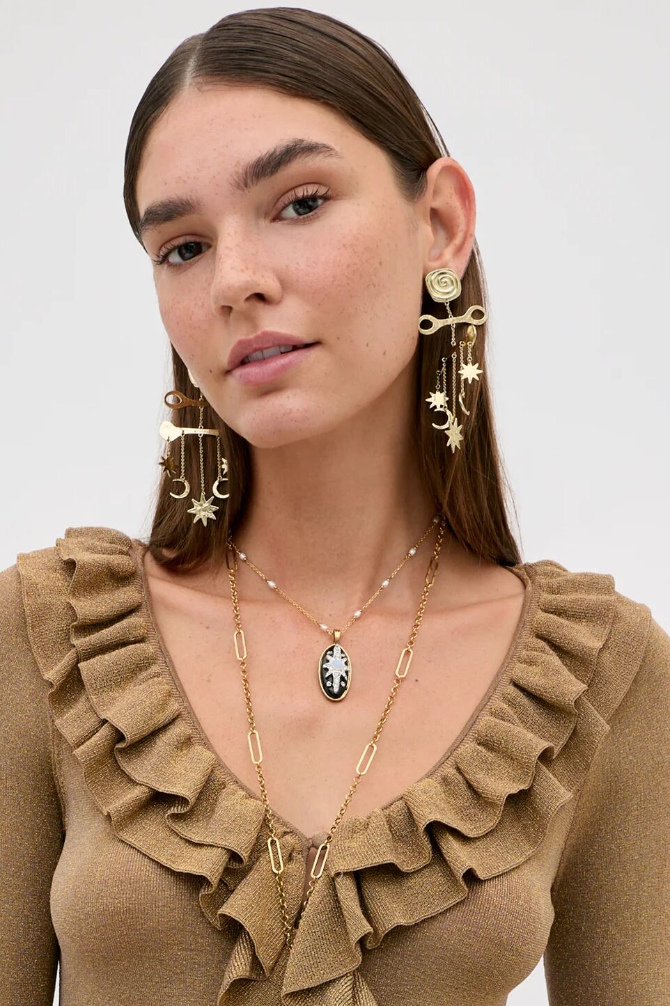 Model wearing Shay - Gold earrings featuring sun and moon charms, a layered gold chain necklace, and a small black oval pendant with a starburst design.