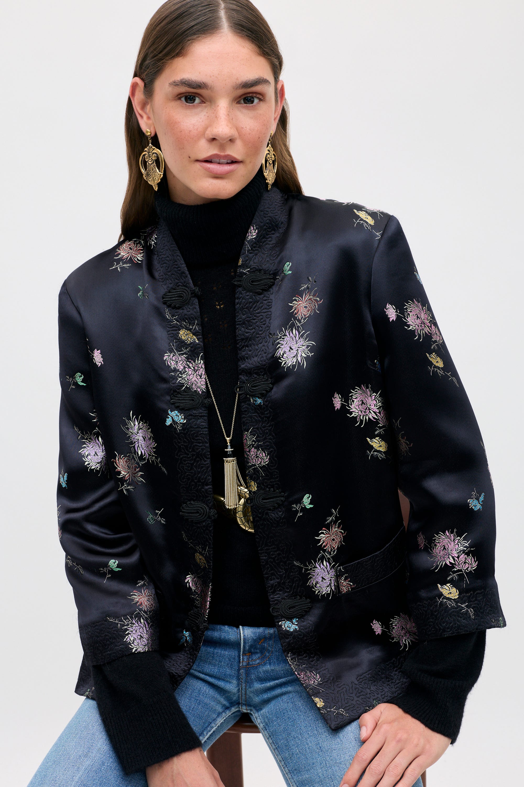 The Sloane - Oriental Flora Black jacket, featuring a floral print, traditional frog closures, and a mandarin collar, worn over a black sweater and jeans.