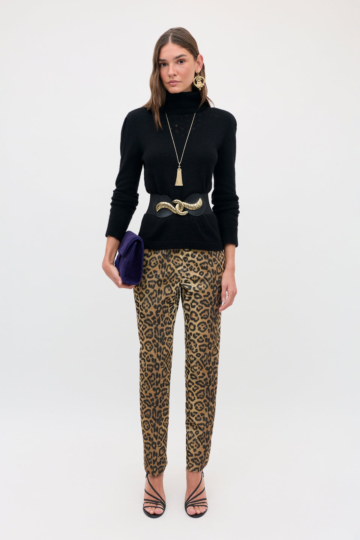 A woman wearing a black turtleneck sweater, a wide black belt with a gold buckle, and Sole - Savannah Leopard Brown trousers, holding a dark blue clutch.