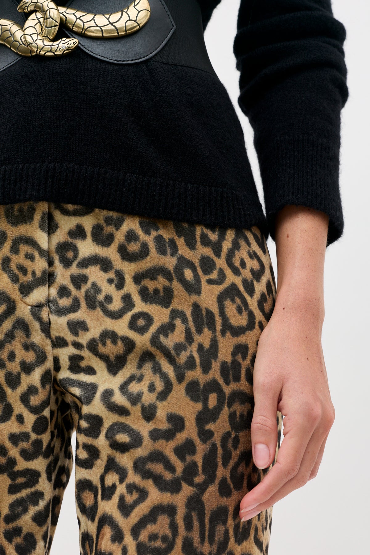 Person wearing a black sweater with a gold snake-shaped belt buckle over Sole - Savannah Leopard Brown pants.