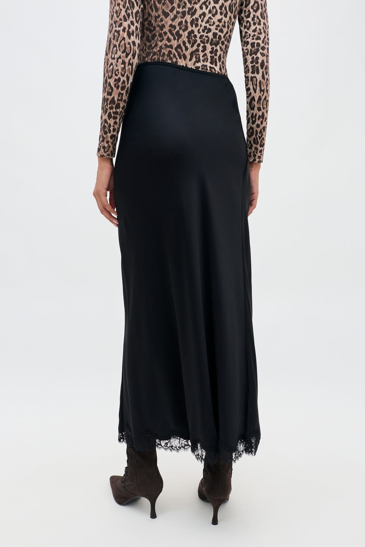 Wearing the Stassie - Black long matte satin skirt with Paris lace trim at the hem, paired with a leopard print long-sleeve top and brown heeled boots, a person stands against a plain light background.