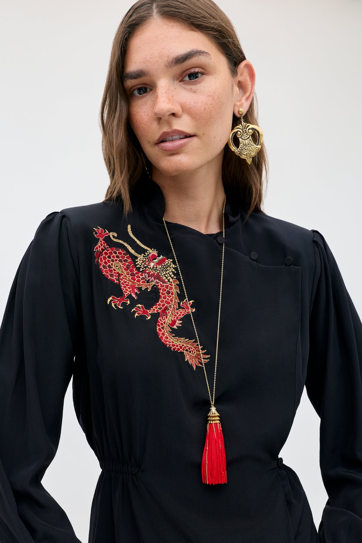 A woman wears the Sylvie Rose - Dragon Embroidery Red, a black long-sleeved dress featuring a red dragon embroidery on the chest and a gold tassel pendant necklace.