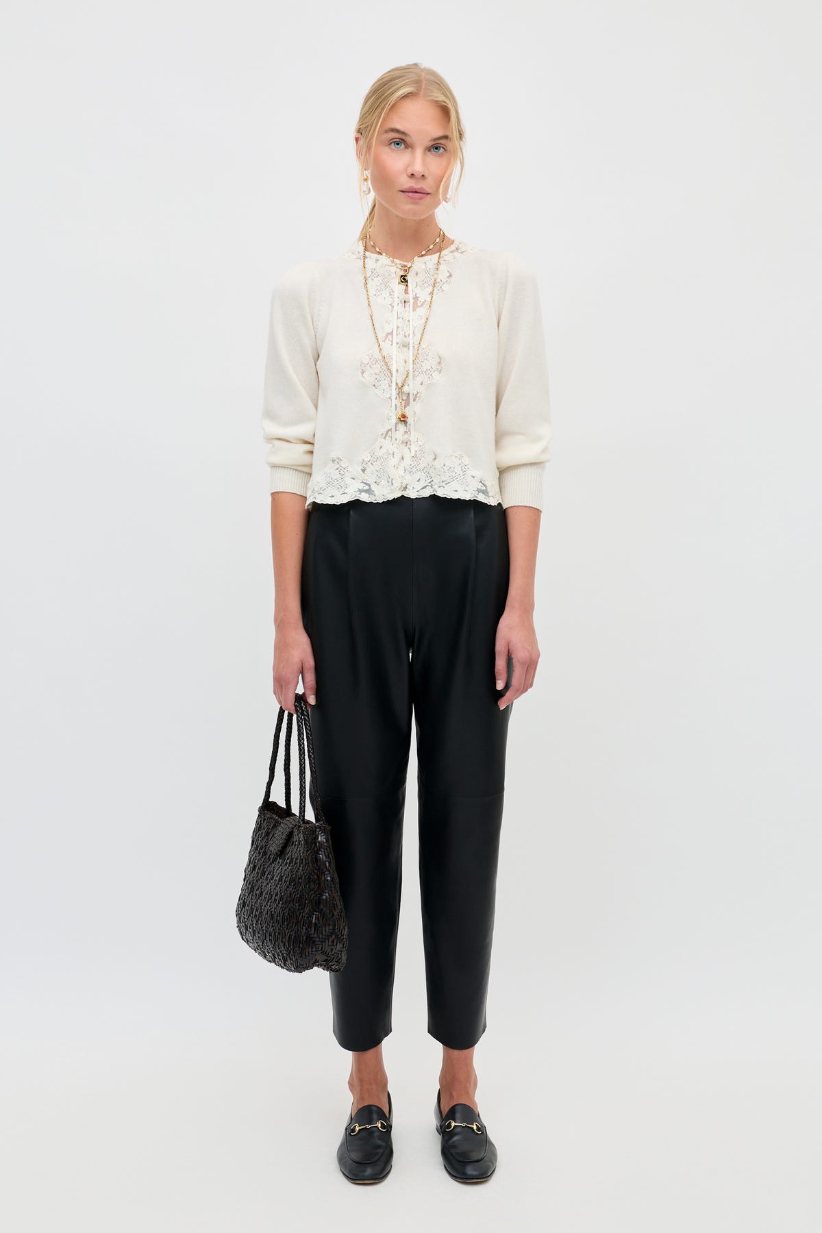 Woman standing against a white background wearing the Tassia - Cream sweater with lace details, paired with black ankle-length pants, black loafers, and holding a textured black handbag. She has blonde hair pulled back and a neutral expression.