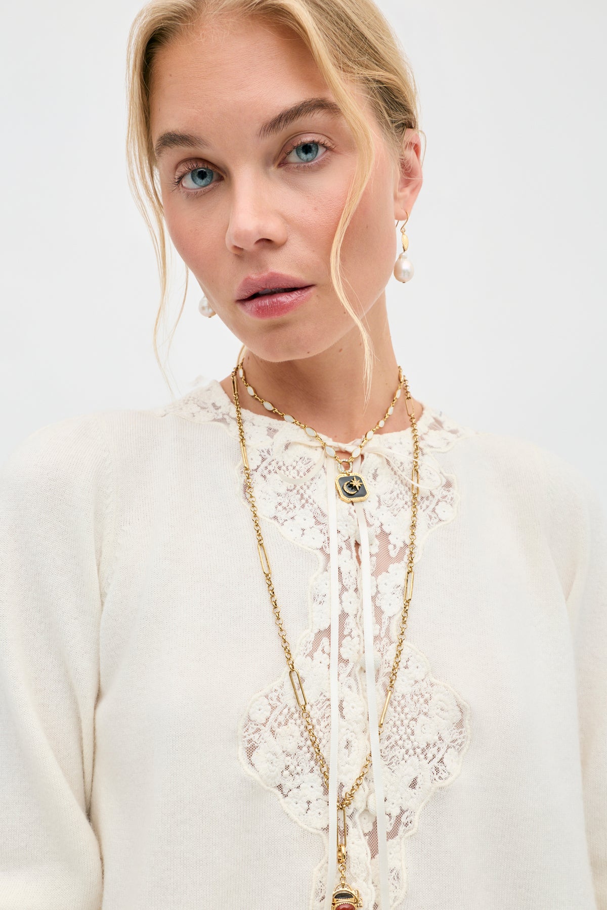 Wearing the Tassia - Cream top with delicate lace trim, a woman accessorized with gold necklaces and pearl earrings gazes at the camera against a plain light background.