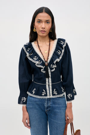 A woman is wearing the Solene Top Flower Trail Embroidery Navy—a navy cotton blouse adorned with white floral embroidery, delicate lace trim, and long sleeves—paired with blue jeans. The vintage-inspired top features a ruffled V-neck that lends an elegant touch to her look. She has long dark hair, gold earrings, layered necklaces, and carries a brown bag while standing against a plain light background.