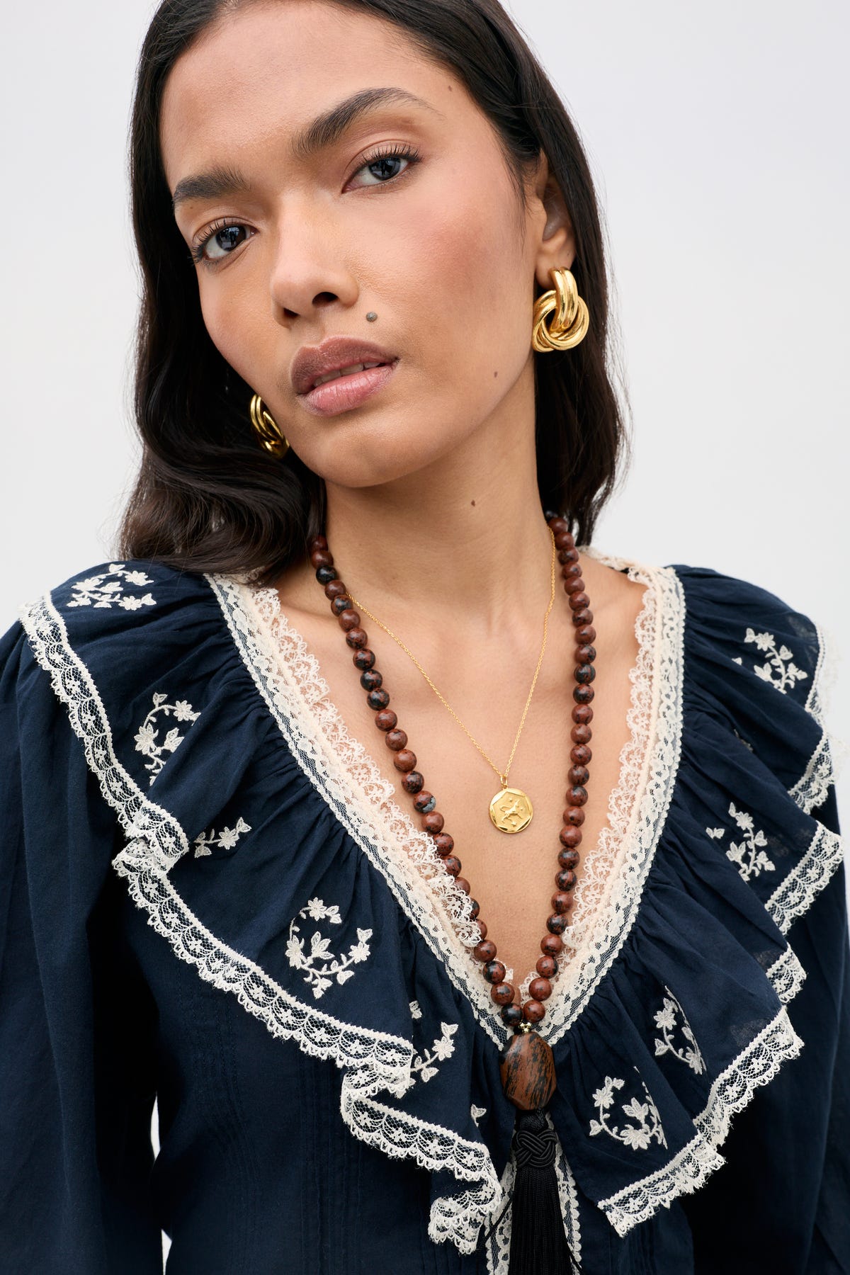 A woman with long dark hair wears the solene top flower trail embroidery navy, featuring ruffles and lace details, paired with gold hoop earrings and layered necklaces. She gazes at the camera with a neutral expression against a simple light background.