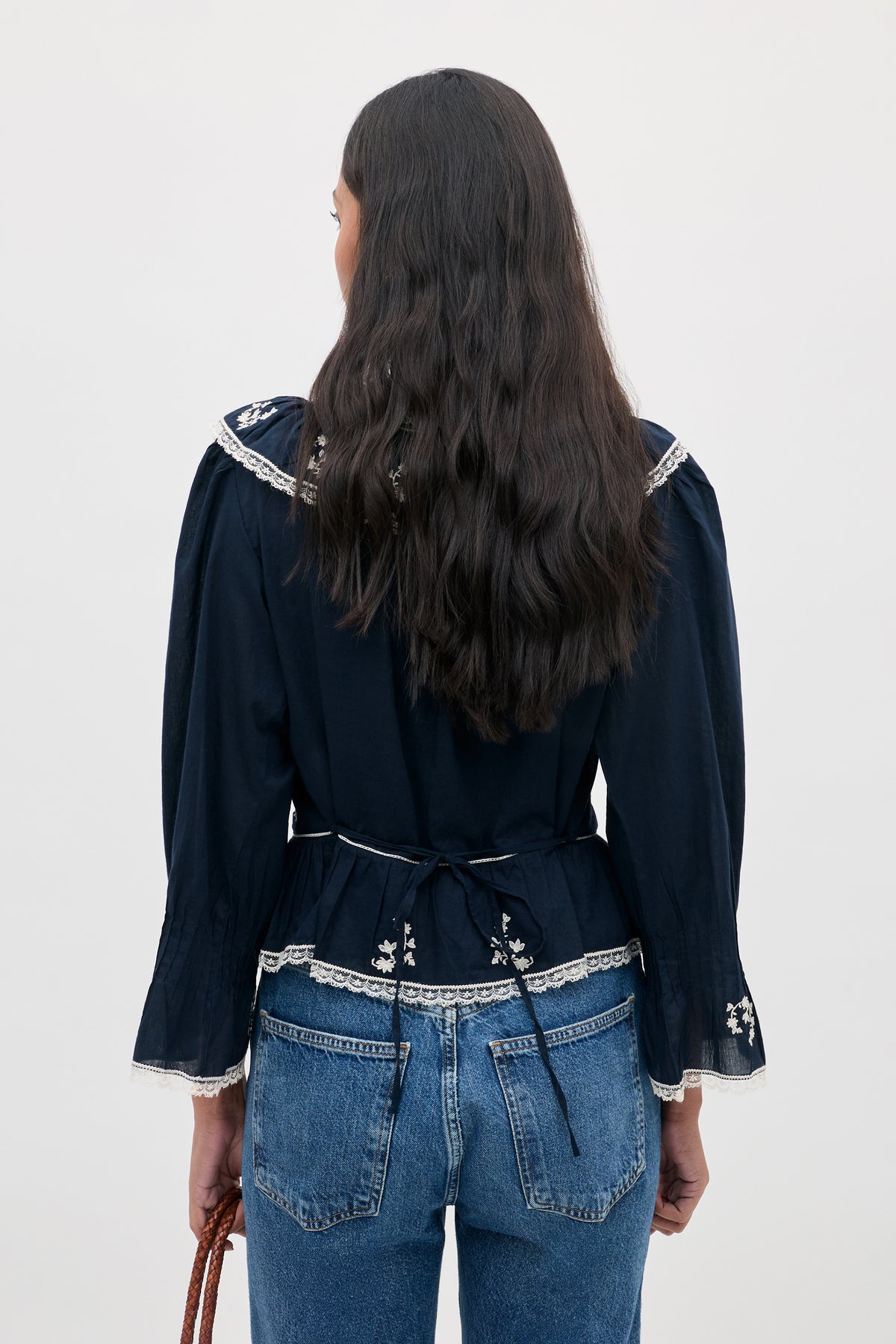 A woman with long wavy hair, shown from the back against a plain light background, is wearing the Solene Top Flower Trail Embroidery in navy paired with blue jeans.
