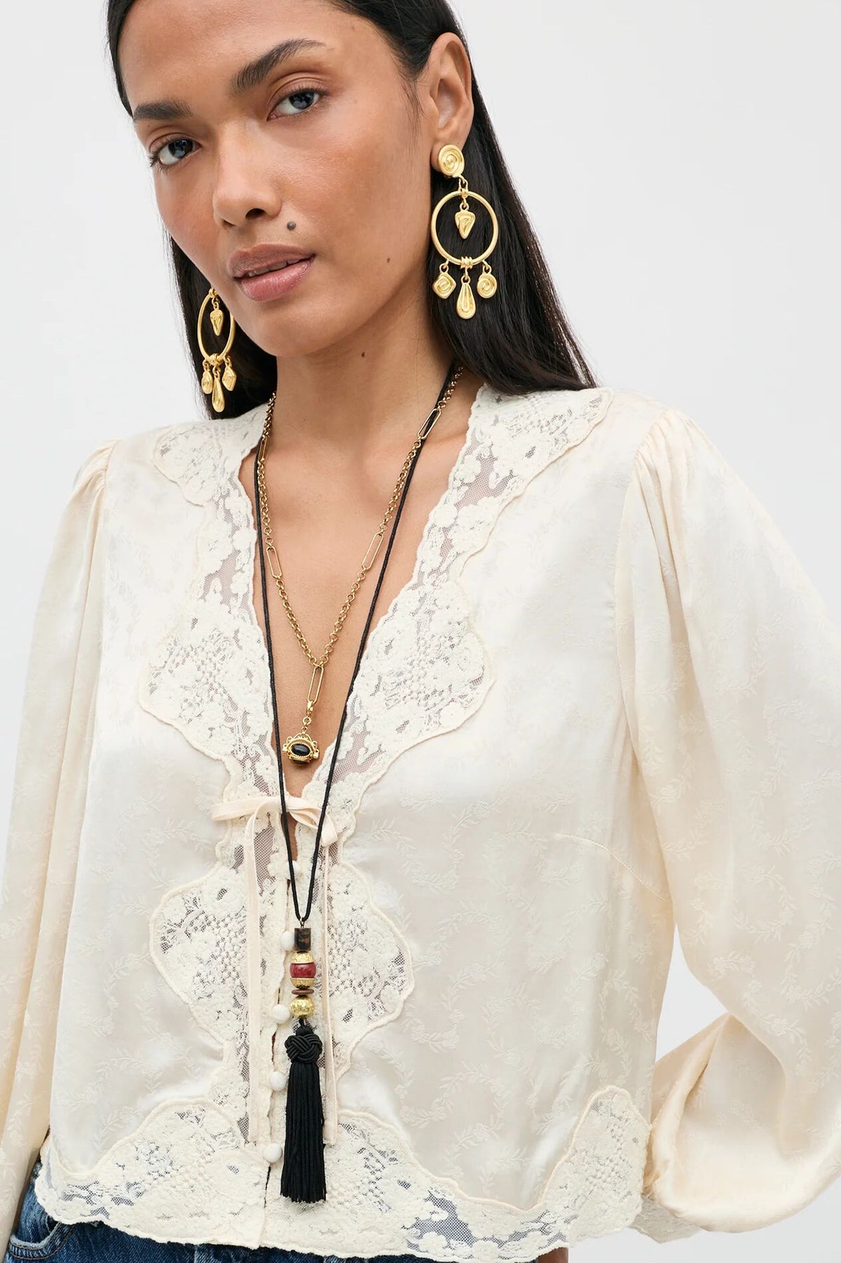A woman wears the Rossa - Black blouse, a cream-colored, long-sleeved lace top, gold hoop earrings with hanging pendants, and a layered pendant necklace with a black tassel.
