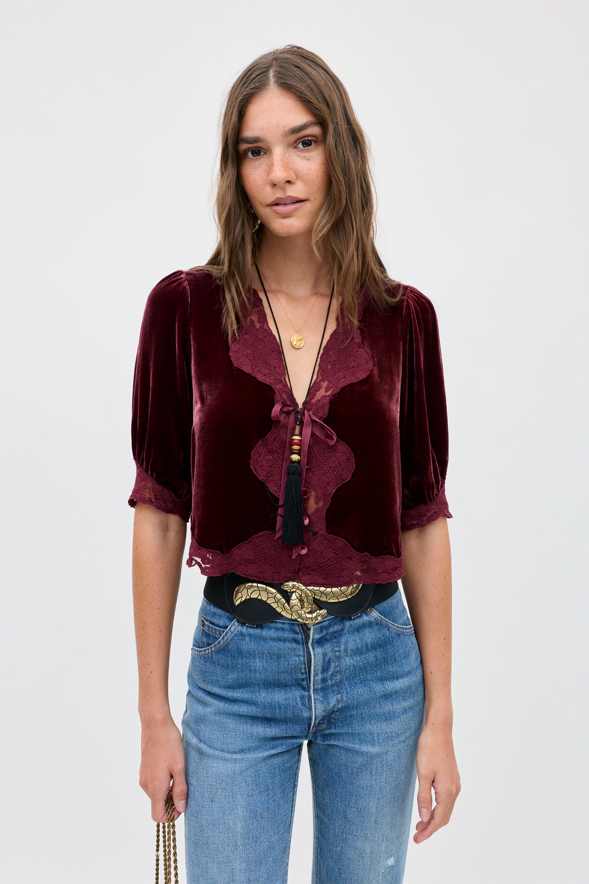 A person wearing an Ursula - Bordeaux short-sleeved, burgundy silk-velvet top with lace trim, a long tassel pendant necklace, and a black belt with a gold-tone snake buckle over blue jeans.