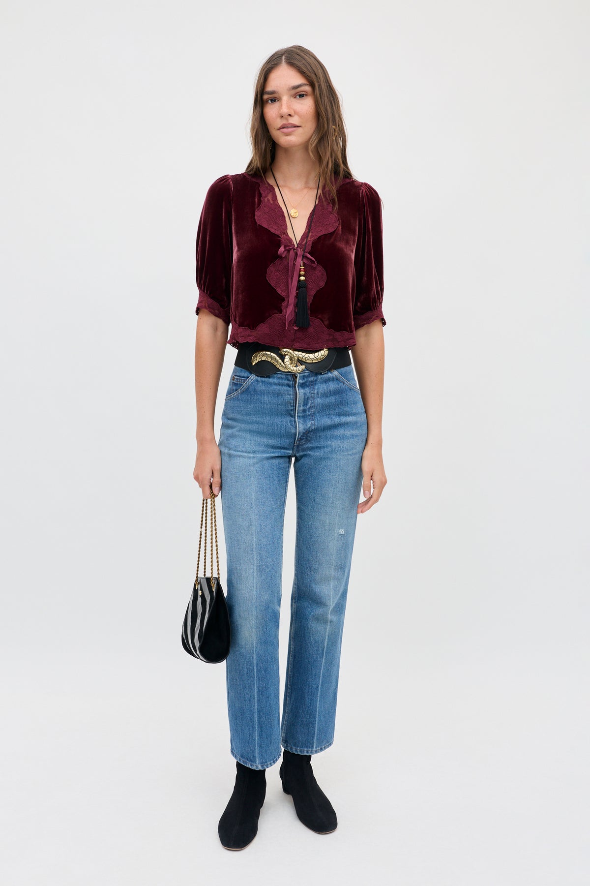 A woman wears a vintage-inspired dark red silk-velvet Ursula - Bordeaux blouse with a deep V-neck and lace trim, blue jeans, a black belt with a gold buckle, and black boots while holding a small black handbag.