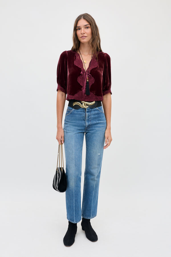 A woman wears a vintage-inspired dark red silk-velvet Ursula - Bordeaux blouse with a deep V-neck and lace trim, blue jeans, a black belt with a gold buckle, and black boots while holding a small black handbag.