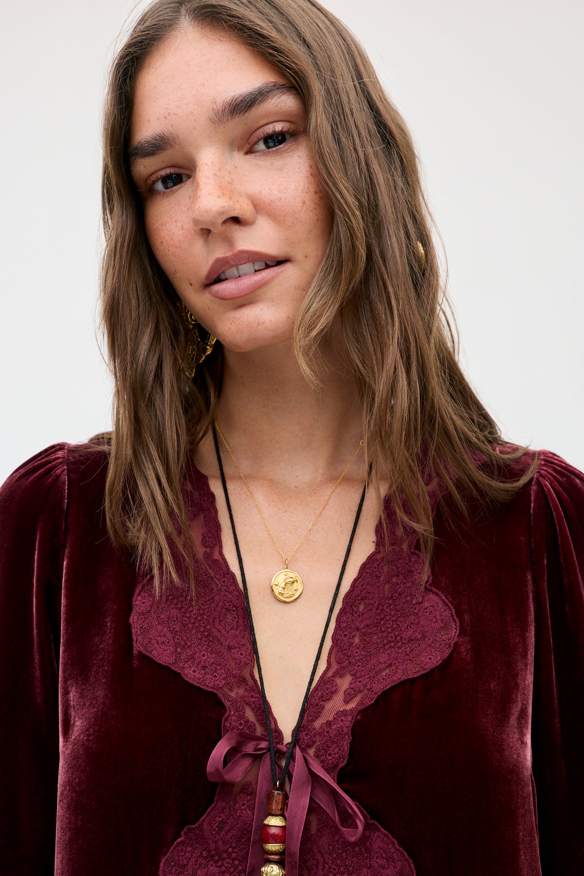 A woman wears the Ursula - Bordeaux top, a vintage-inspired dark red silk-velvet design with lace trim, styled with layered gold and beaded necklaces.