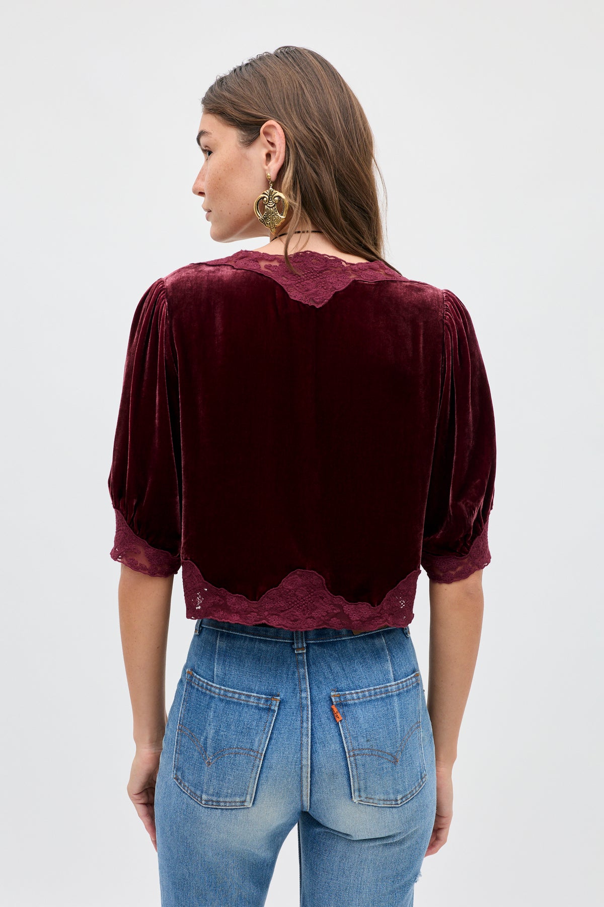 A person seen from the back wearing the Ursula - Bordeaux, a vintage-inspired dark red silk-velvet top with short puff sleeves and lace trim, paired with blue jeans.