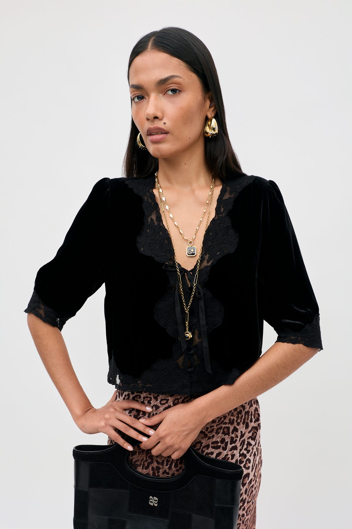 Dressed in the Ursula - Black vintage party top crafted from luxurious black silk velvet with delicate lace trim, a woman stands confidently against a plain background. She completes her look with layered gold necklaces, hoop earrings, a leopard print skirt, and a black handbag as she gazes directly at the camera.