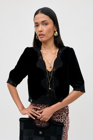 Dressed in the Ursula - Black vintage party top crafted from luxurious black silk velvet with delicate lace trim, a woman stands confidently against a plain background. She completes her look with layered gold necklaces, hoop earrings, a leopard print skirt, and a black handbag as she gazes directly at the camera.