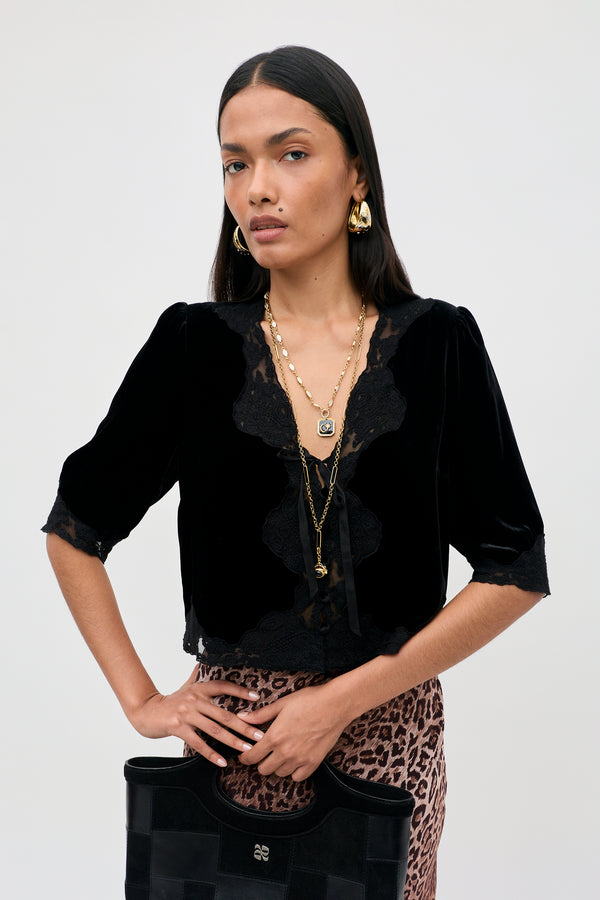 Dressed in the Ursula - Black vintage party top crafted from luxurious black silk velvet with delicate lace trim, a woman stands confidently against a plain background. She completes her look with layered gold necklaces, hoop earrings, a leopard print skirt, and a black handbag as she gazes directly at the camera.