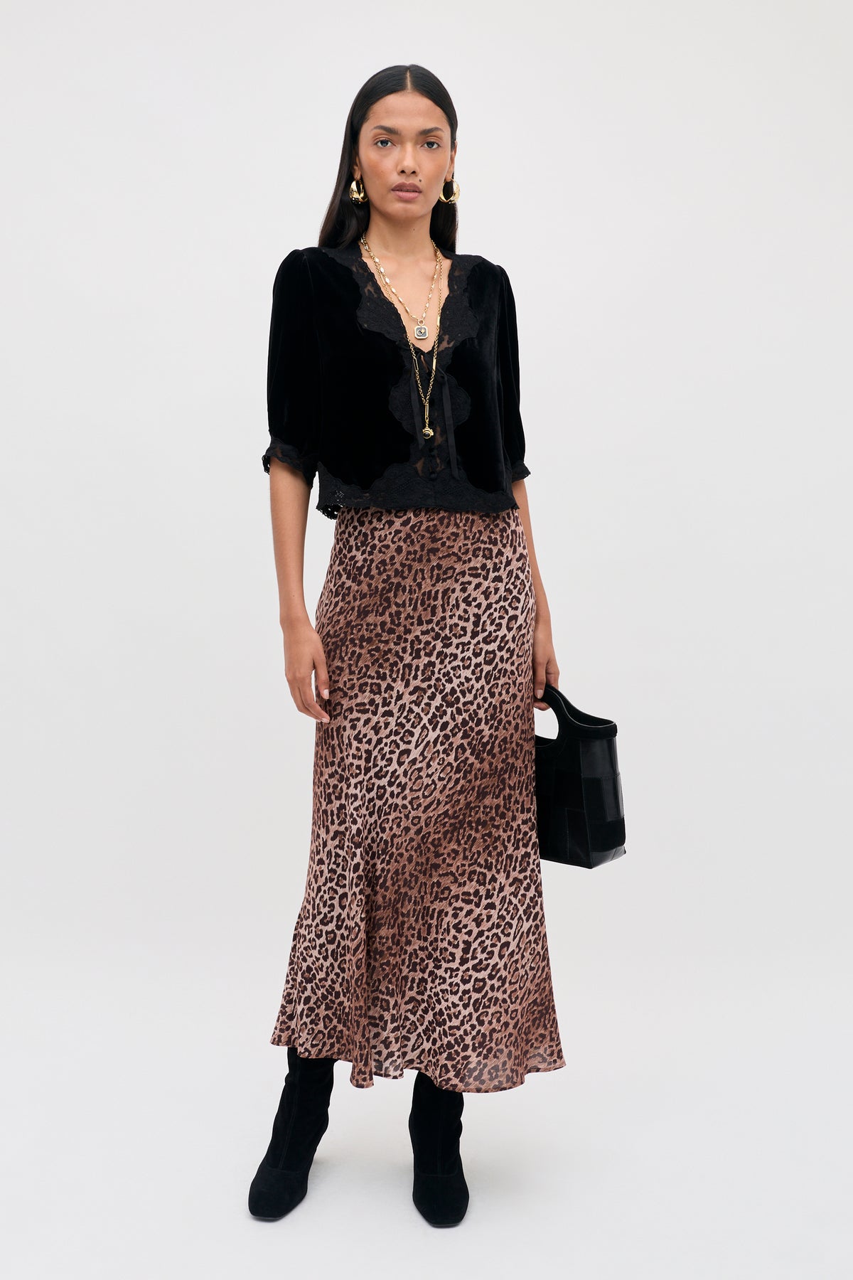 A woman standing against a plain white background wears a vintage party top with lace trim, a leopard print midi skirt, black boots, layered necklaces, hoop earrings, and holds the Ursula - Black handbag.